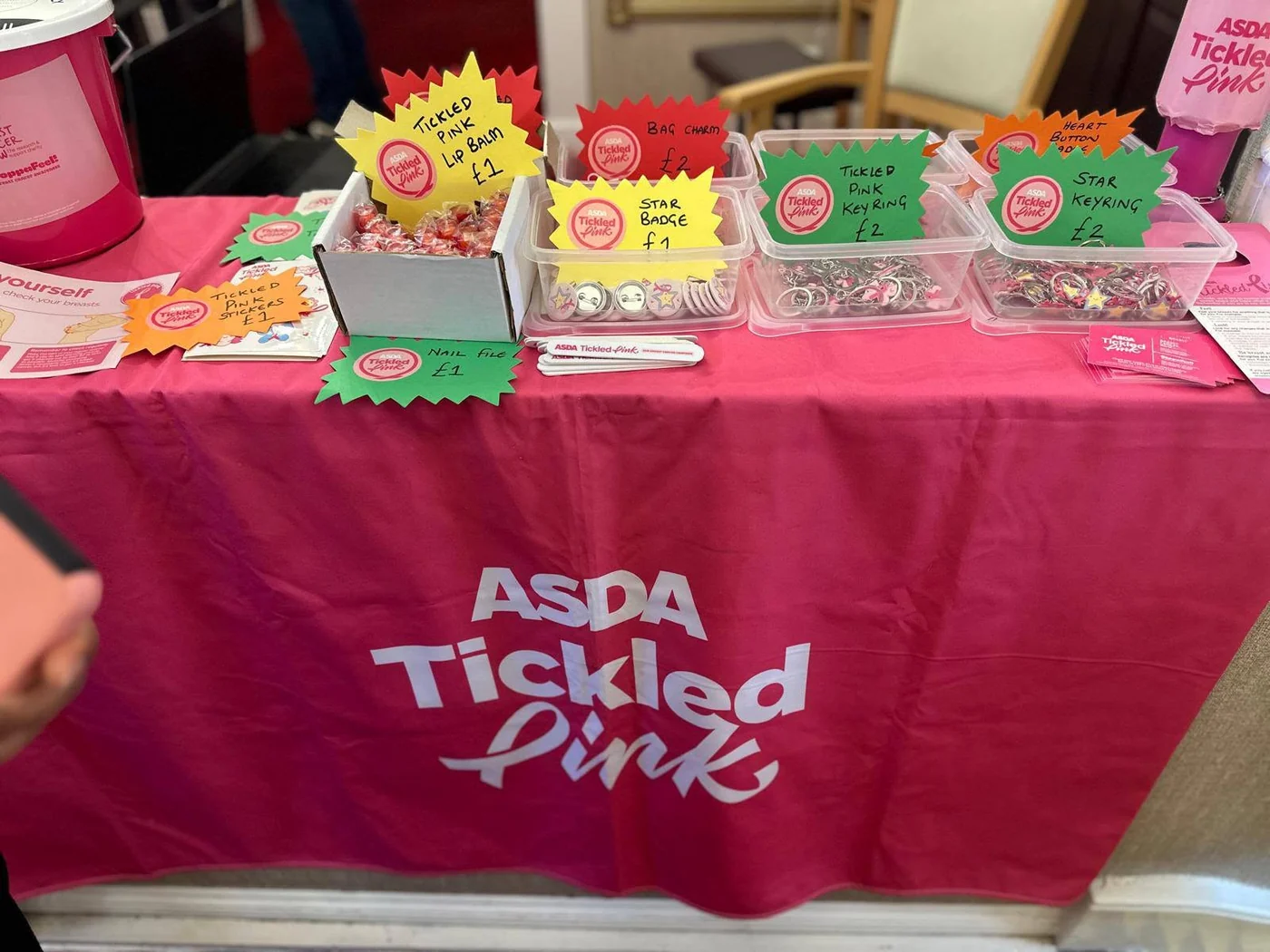 A stall covered with a pink Asda Tickled Pink cloth, laid out with lip balms, badges, keyrings, nail files and stickers, a collection bucket and breast checking leaflets.
